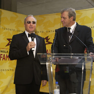 Lorne Michaels with sunglasses and tuxedo, speaking at podium
